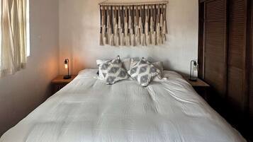 A bedroom with a bed, a wall-mounted macrame piece, a bedside lamp, and a hanging light fixture.
