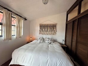 Ein Schlafzimmer mit einem großen Bett, einer Wanddekoration aus Makramee, einer Nachttischlampe und einem Fenster mit Vorhängen.