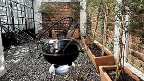 A barbecue grill in a garden area with a brick wall, a small tree, and metal chairs.