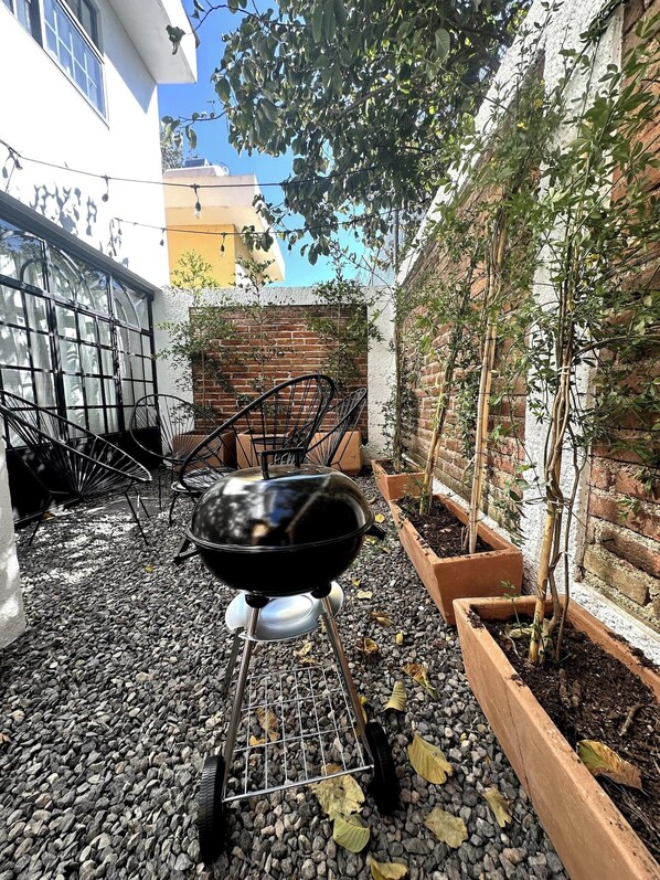 Un barbecue installé dans un coin du jardin, avec un mur en briques, un petit arbre et des chaises en métal.