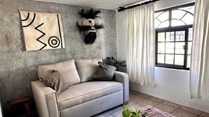 A living room with a sofa, a wooden coffee table with a plant, a window with curtains, and a wall with a geometric artwork.