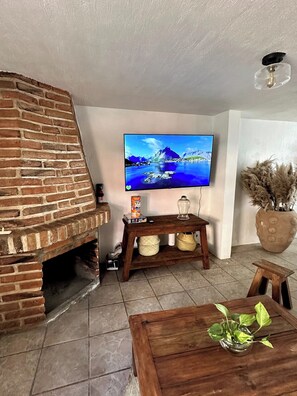 Un salon avec une cheminée en briques, un téléviseur fixé au mur, une table basse en bois et une plante en pot.