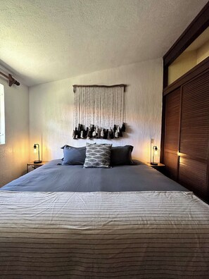 A bedroom with a large bed, two bedside tables with lamps, a wall-mounted macrame piece, and a wooden wardrobe.