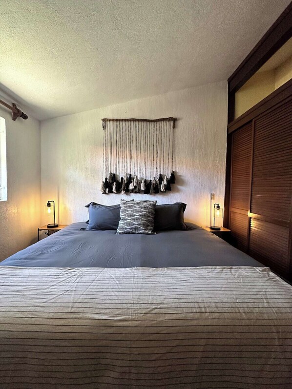Une chambre avec un grand lit, deux tables de chevet avec des lampes, une décoration murale en macramé et une armoire en bois.