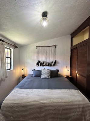 Un dormitorio con una cama grande, dos mesitas de noche con lámparas, un adorno de macramé en la pared y un armario de madera.