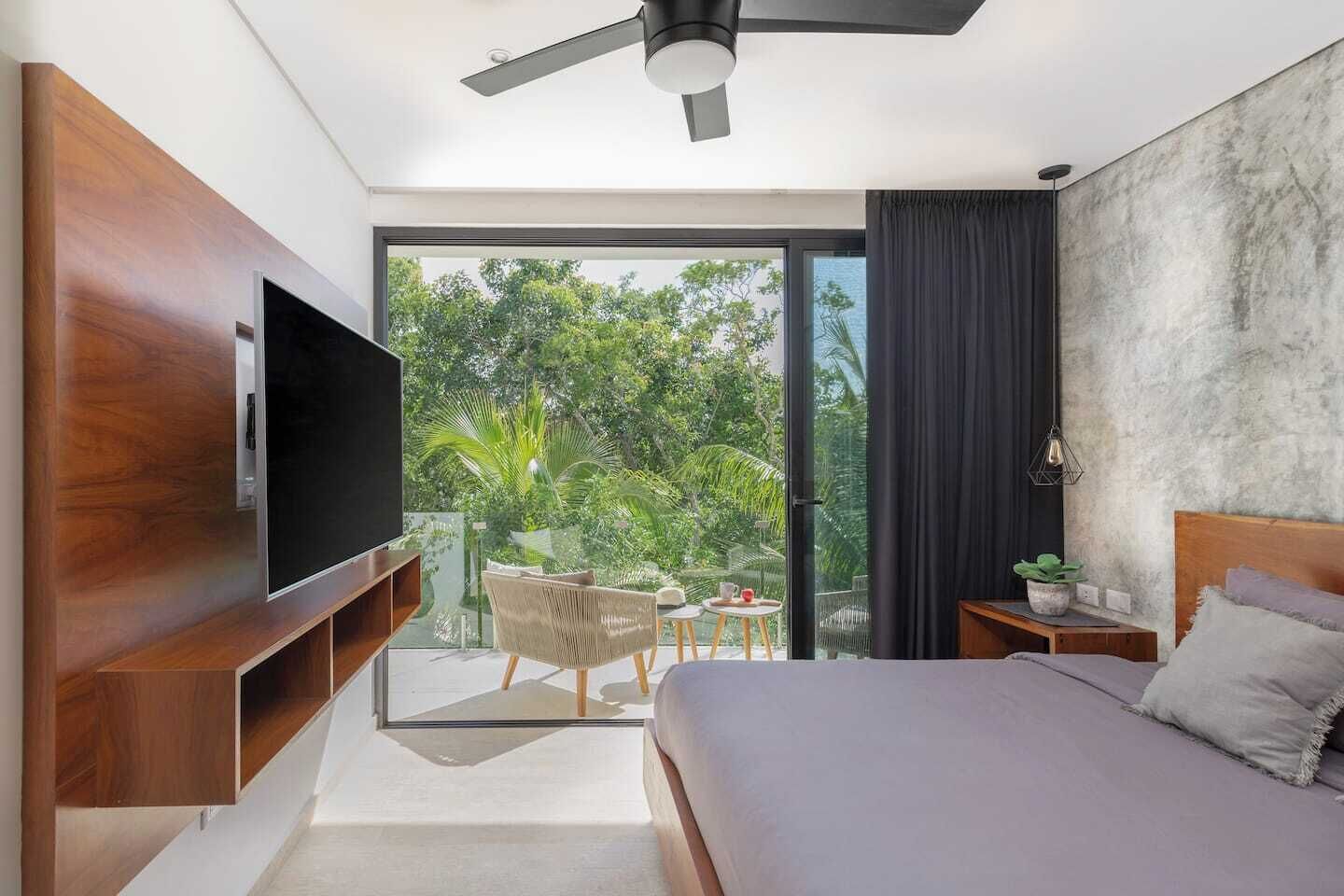 Une chambre moderne dotée d’un grand lit, d’un téléviseur à écran plat, d’un ventilateur de plafond et d’un balcon aménagé avec du mobilier d’extérieur.