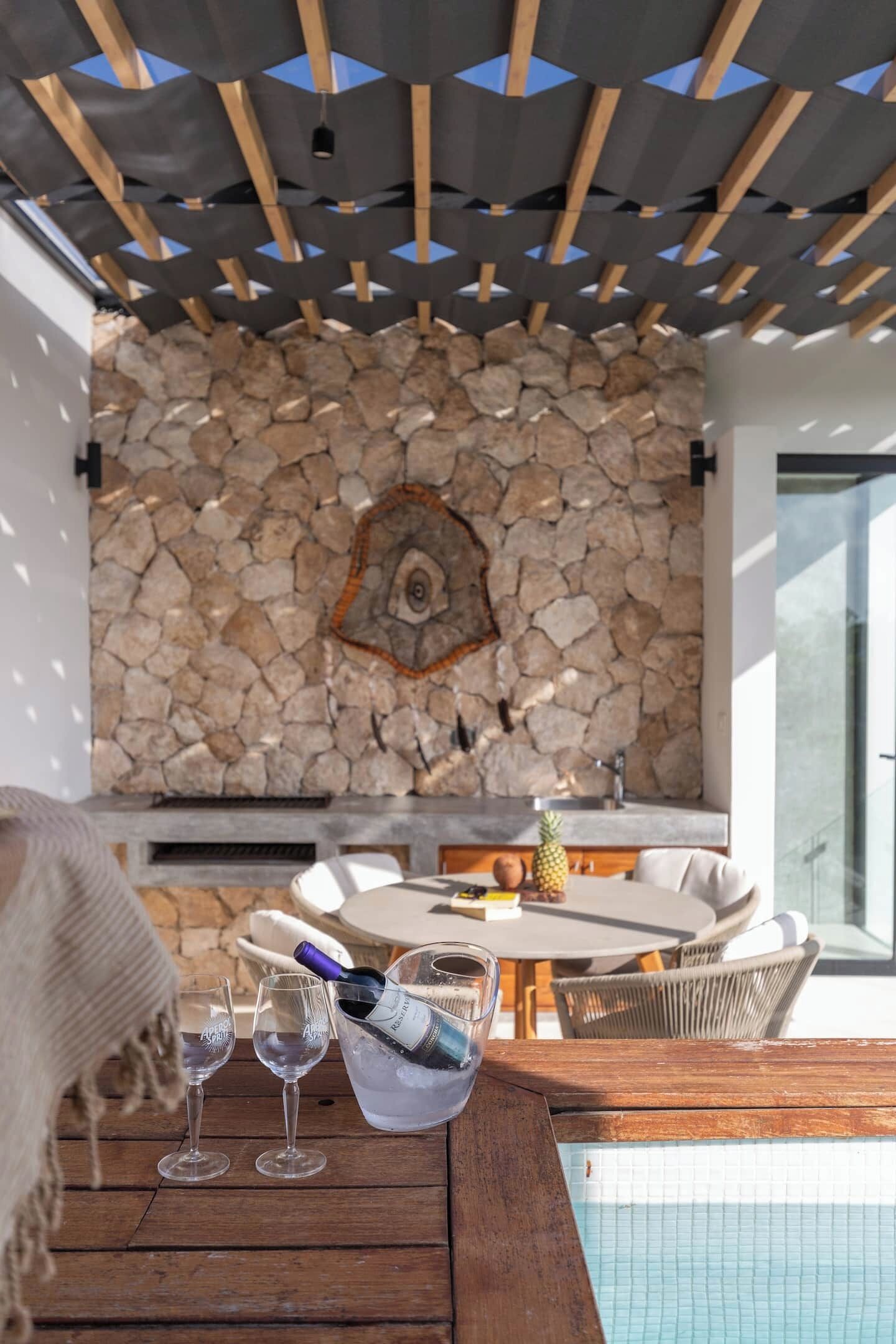 A poolside area with a stone wall, a wooden table with a bottle of champagne, and two glasses.
