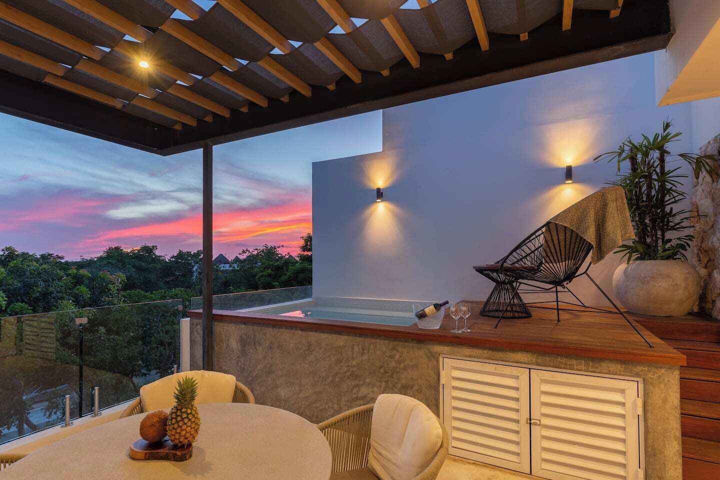 A modern outdoor patio with a table, chairs, a wooden ceiling, and a view of the sunset.