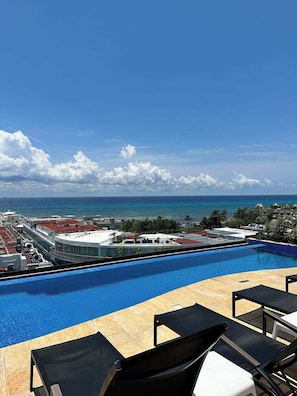 Ein Pool mit Liegestühlen, Blick auf das Meer und Gebäude im Hintergrund.