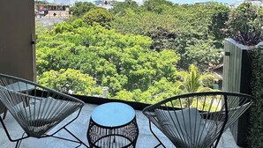 Ein Balkon mit zwei Stuhlen aus Drahtgitter und einem kleinen runden Tisch, mit Blick auf eine üppige grüne Landschaft und entfernte Gebäude.