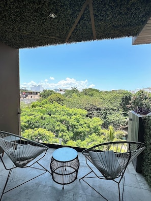 Ein Balkon mit zwei Stuhlen aus Drahtgitter und einem kleinen runden Tisch, mit Blick auf eine üppige grüne Landschaft und entfernte Gebäude.