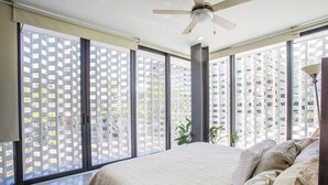 Un dormitorio con cama, ventilador de techo y ventanas amplias con vistas a edificios altos.