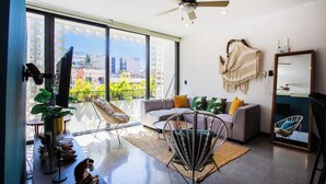 Una sala de estar moderna con sofá, sillones, ventilador de techo y ventanas amplias con vista a la ciudad.