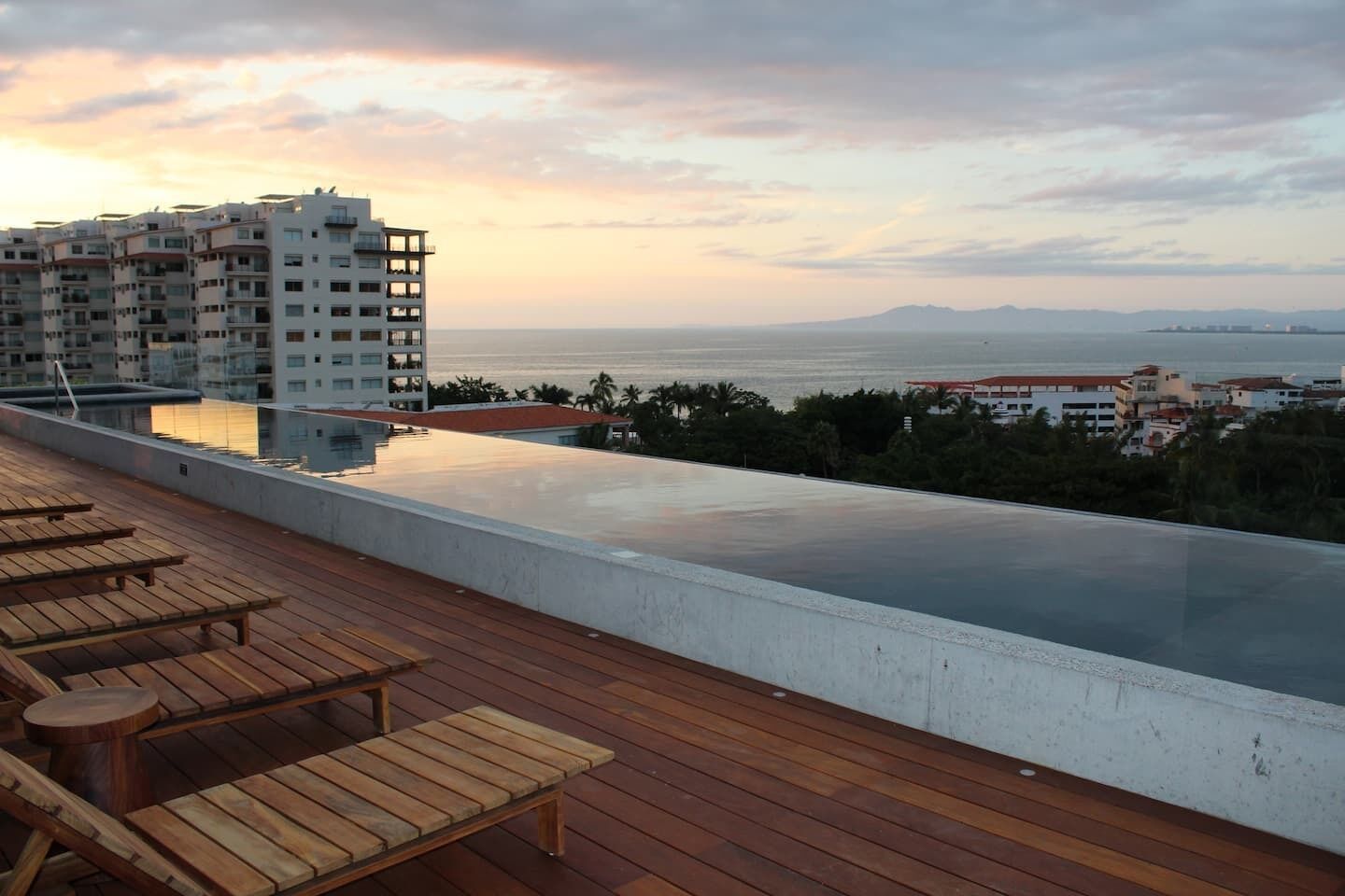 A rooftop pool with wooden lounge chairs, a view of the sea, and a high-rise building in the background.