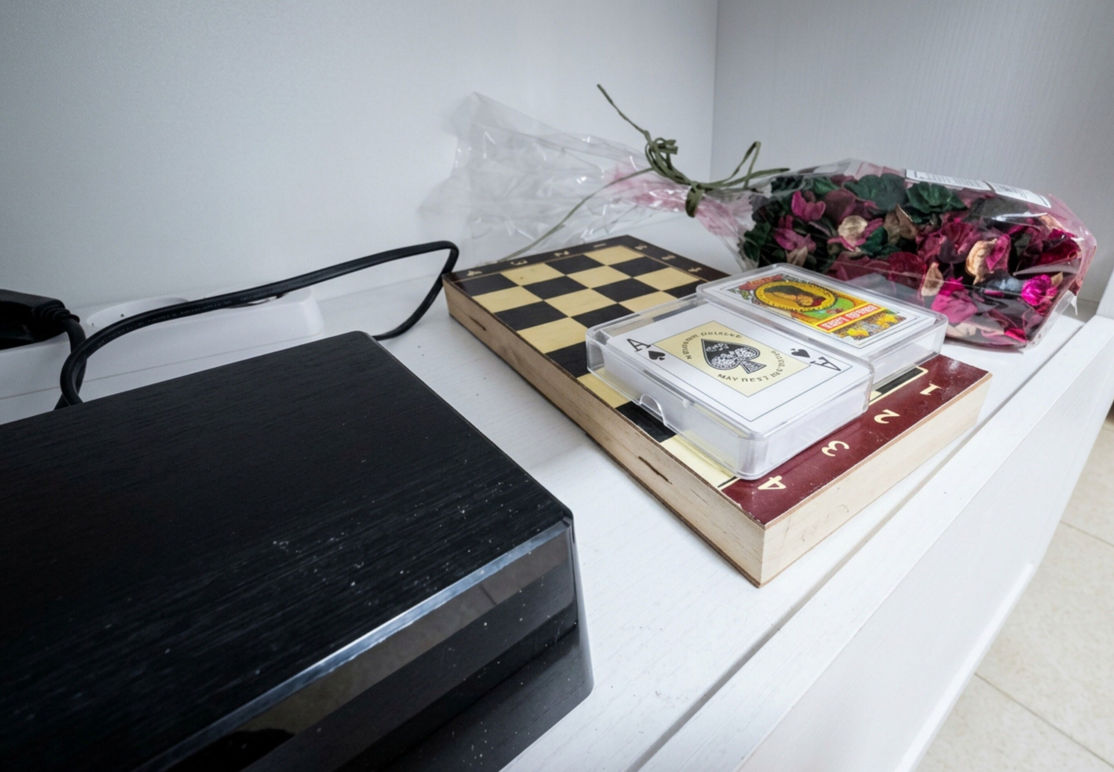 A desk with a black electronic device, a chessboard, a deck of cards, and a bouquet of flowers.