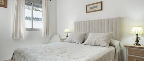 A bedroom with a bed, bedside lamps, a nightstand, a chair, and a window with curtains.