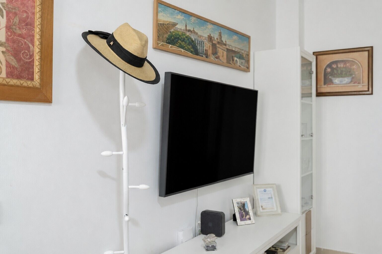 A room with a mounted flat-screen TV, a hat on a stand, and framed artwork on the walls.