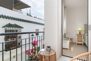 Un balcón con plantas en macetas, una mesa de mimbre y vistas a un edificio blanco con tejado de azulejos.