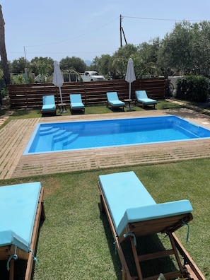 A rectangular swimming pool with a wooden deck surrounding it, equipped with lounge chairs and umbrellas.