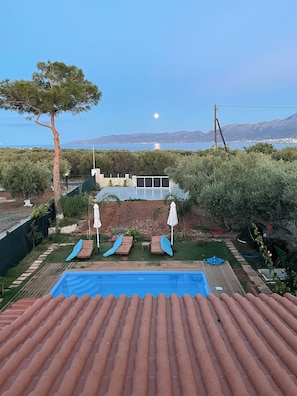 A swimming pool with lounge chairs and umbrellas, a view of the sea, and a mountainous landscape in the background.