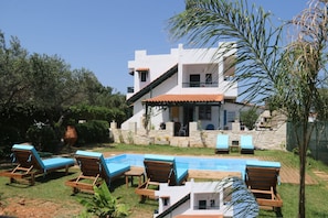 A modern two-story house with a swimming pool, lounge chairs, and a stone wall.