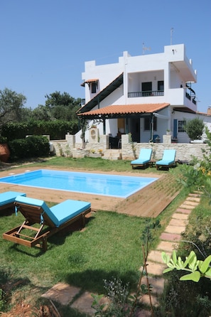 A modern two-story house with a swimming pool, two lounge chairs, and a stone wall.