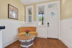 Ein Zimmer mit gelber Wand, weißem Zierleisten, Holzboden, einem gelben Stuhl, einem runden Tisch mit Blumenvase und einer weißen Tür mit Fenster.