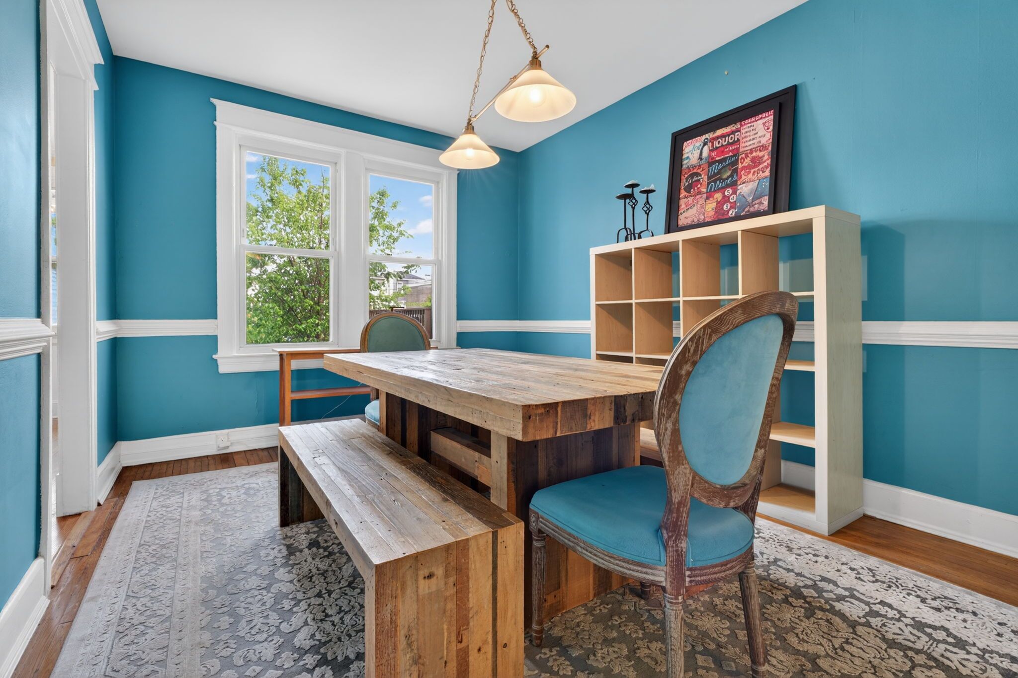 A room with teal walls, a wooden dining table, a bench, a chair with a blue cushion, a window, and a bookshelf.