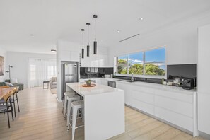 A modern kitchen with white cabinetry, a central island, pendant lighting, and a view of greenery through large windows. - 22 Dent Crescent - Spacious Coastal Retreat with Stunning Ocean Views (Port Macquarie)