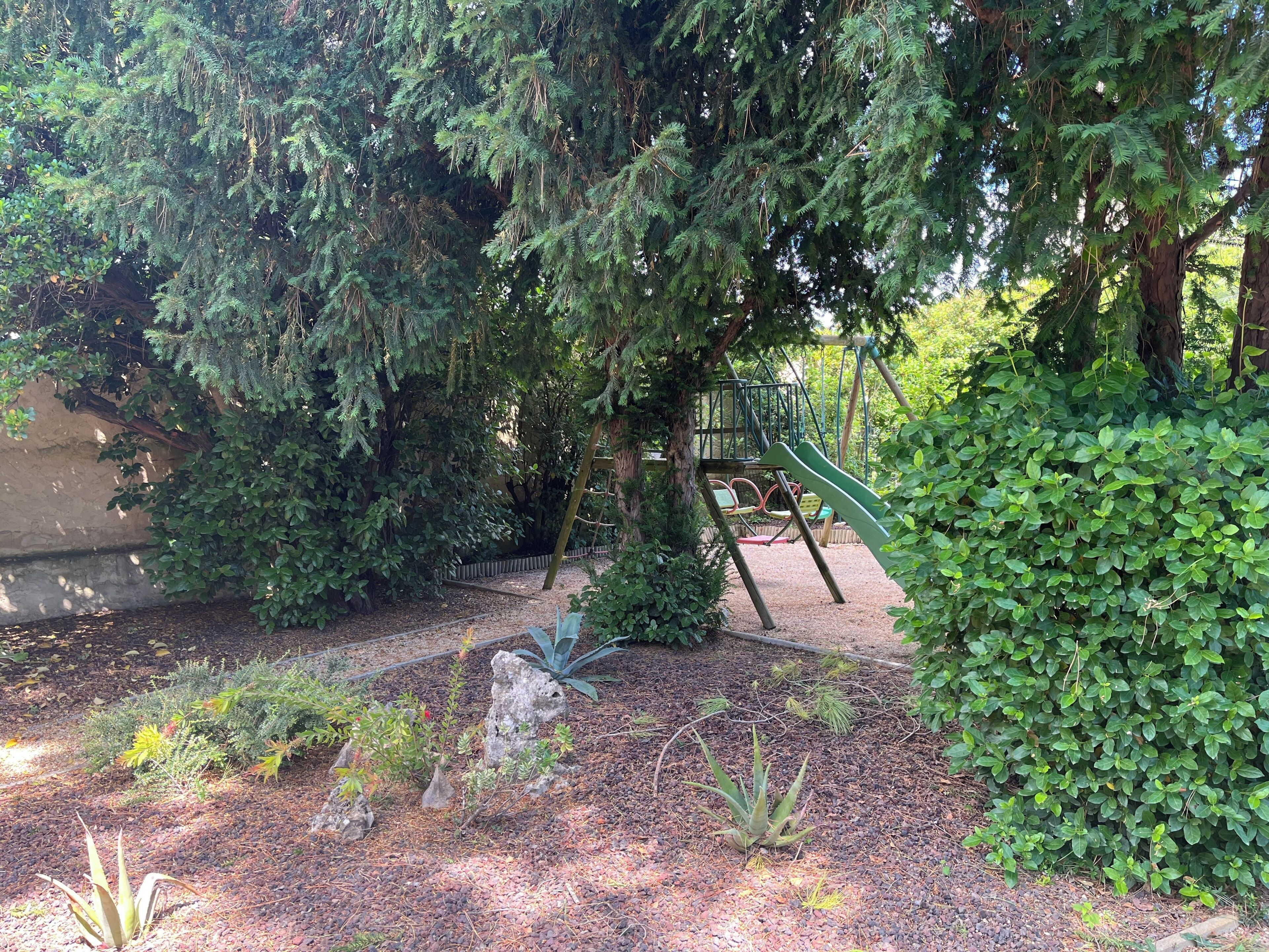 Un parque infantil con tobogán y estructuras para escalar, rodeado de follaje denso y un camino de grava.