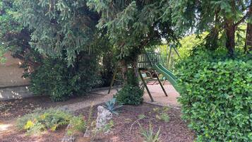 Un parque infantil con tobogán y estructuras para escalar, rodeado de follaje denso y un camino de grava.