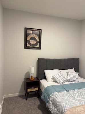 Une chambre à coucher avec un lit, des oreillers, une table de chevet, une lampe et une œuvre d’art encadrée accrochée au mur.