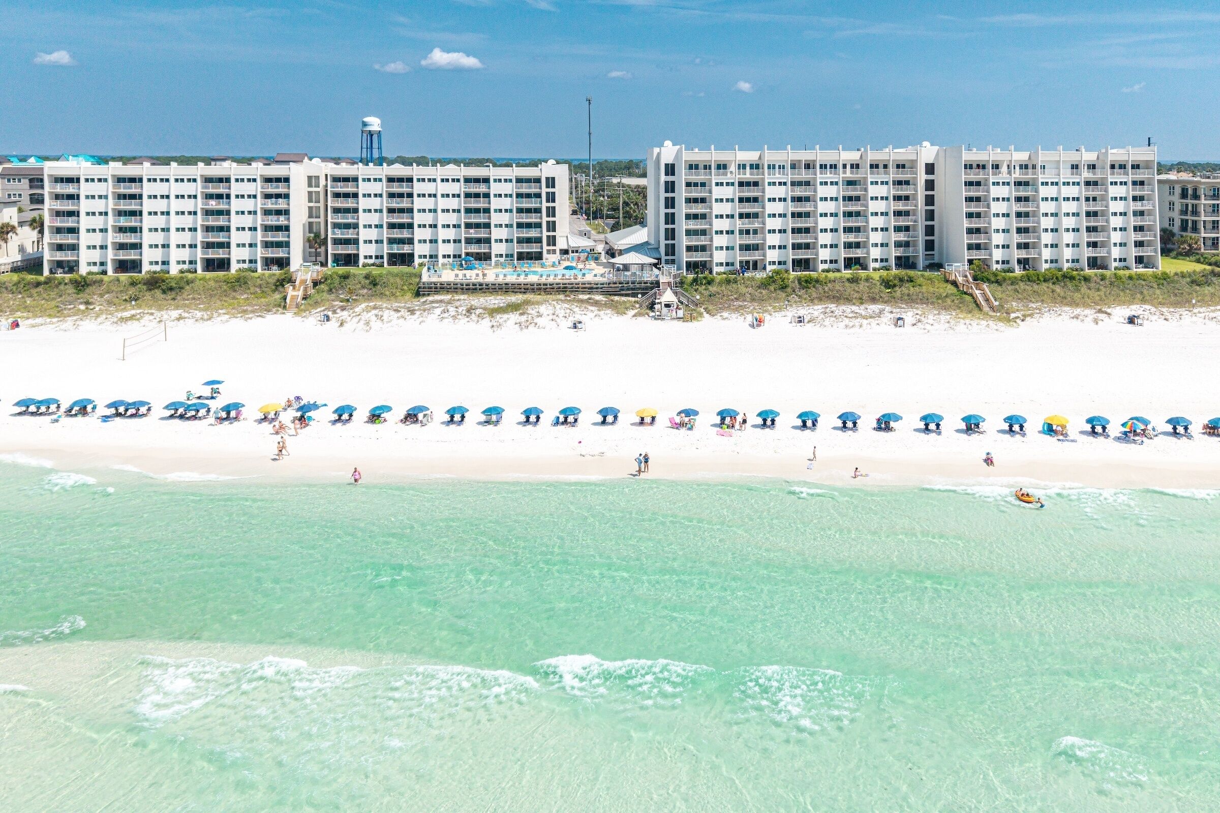 Un hôtel en bord de mer, avec plusieurs étages, une plage de sable et une rangée de parasols.