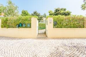 A gated entrance with a cobblestone path, green foliage, and a clear sky. - Vilamoura Garden and Pool Apartment (Vilamoura)