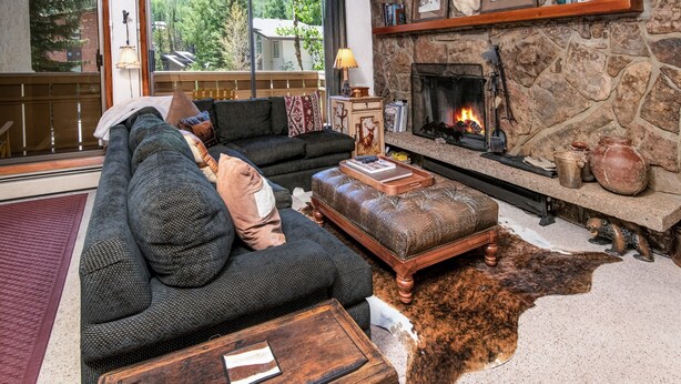 A cozy living room with a stone fireplace, a large sofa, and a leather ottoman. - Scorpio #101 2 Bedroom Condo (Vail)