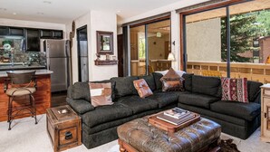 A living room with a sectional sofa, a coffee table, a leather ottoman, a bar area, and a large window with a view of the outdoors. - Scorpio #101 2 Bedroom Condo (Vail)