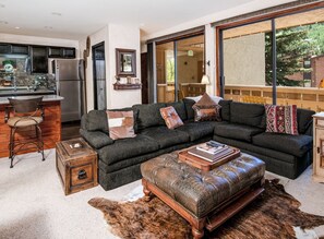 Ein Wohnzimmer mit einem Ecksofa, einem Couchtisch, einem Lederhocker, einer Bar und einem großen Fenster mit Blick ins Freie.