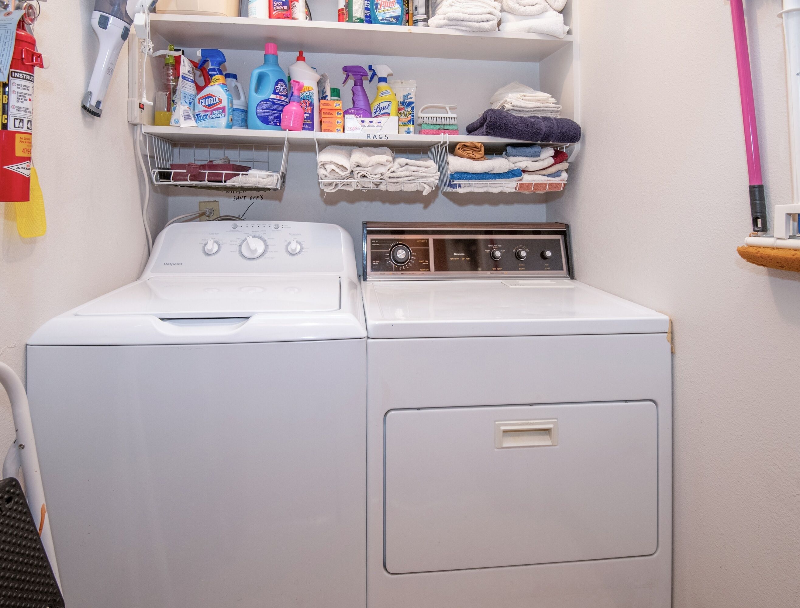 Una lavadora de carga frontal con panel de control, un estante superior con productos de limpieza y toallas dobladas, y un extintor en la pared.