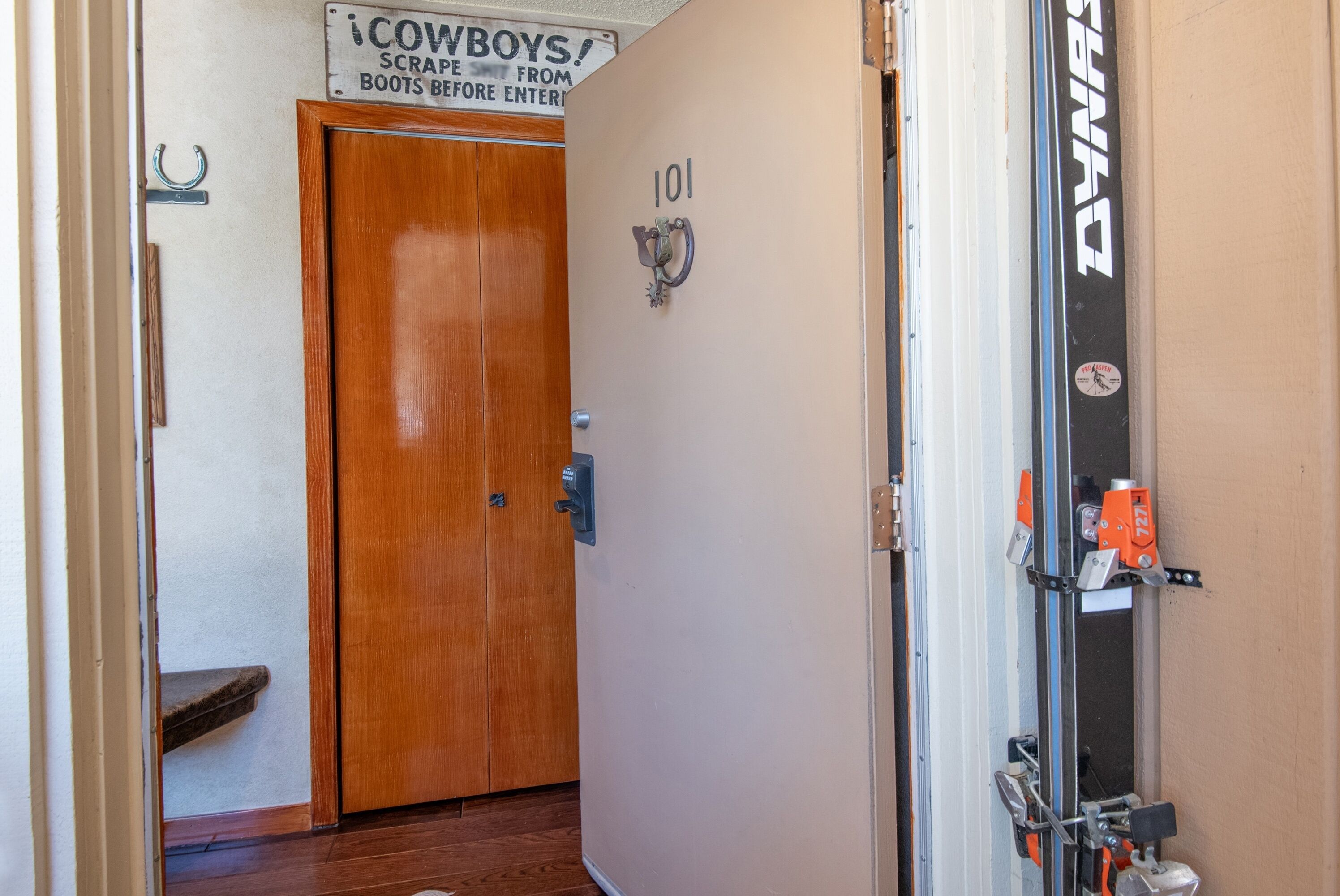 A hallway with a wooden door labeled '101,' a sign above instructing to 'SCRAPE FROM BOOTS BEFORE ENTERING,' and a doorbell with a metal handle.