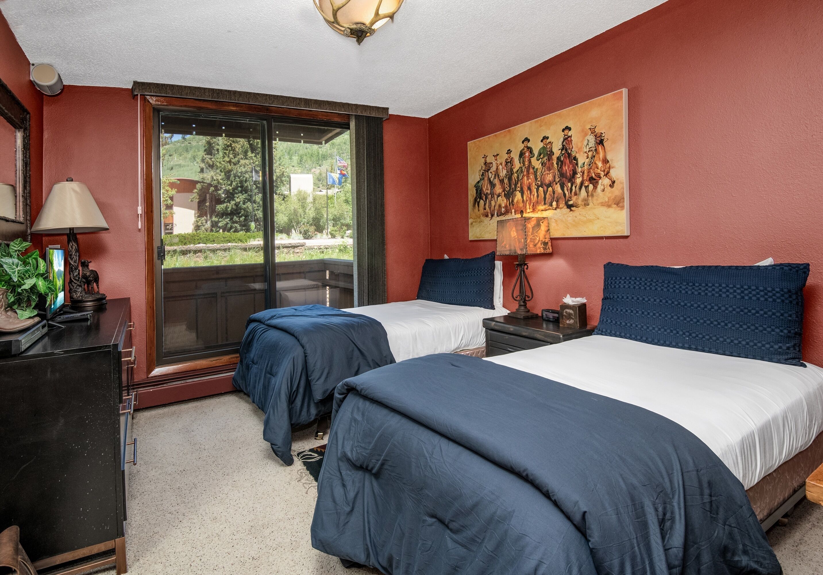 A bedroom with two beds, a painting, a lamp, and a view through a sliding door.