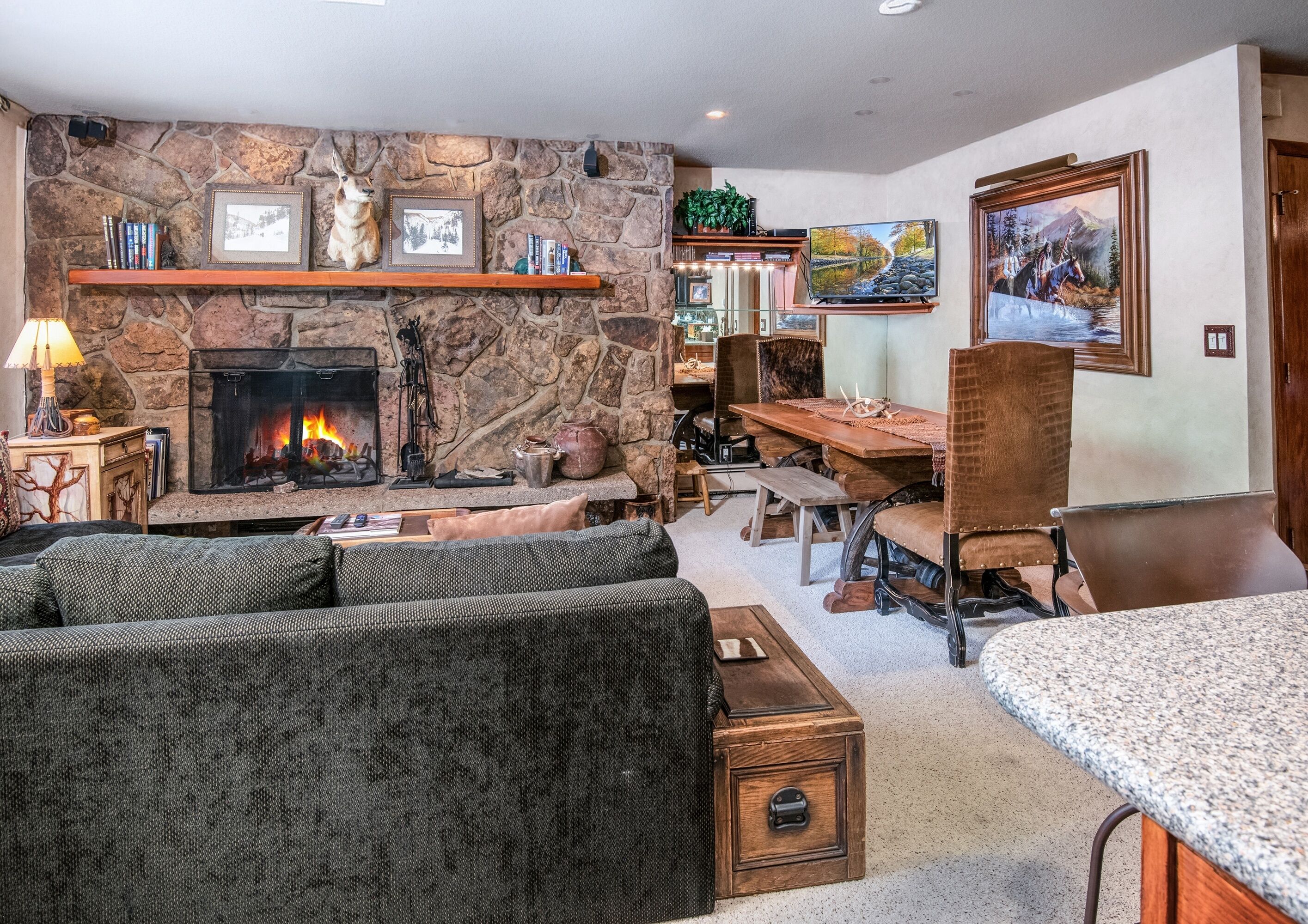 Ein Wohnzimmer mit Kamin aus Stein, einem Essbereich mit Holztisch und -stühlen sowie einem Bücherregal mit diversen Gegenständen.