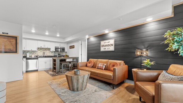 A modern living room with a leather sofa, a coffee table, and a dining area with white cabinets and a kitchen in the background. - Rc305a- The Landing Pad 2 Bedroom Condo (Branson)
