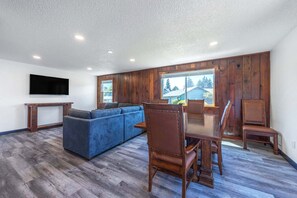 A spacious living room with a large sofa, wooden dining table, and chairs, a flat-screen TV mounted on the wall, and large windows with a view of the outside.