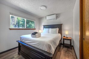 Un dormitorio con cama, mesitas de noche, aire acondicionado y ventana con vista a árboles.