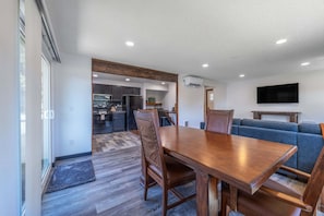 A modern kitchen with built-in appliances, a dining area with a wooden table and chairs, a blue sofa, and a mounted TV. - Tails & Trails 3 Bedroom Home (Portland)