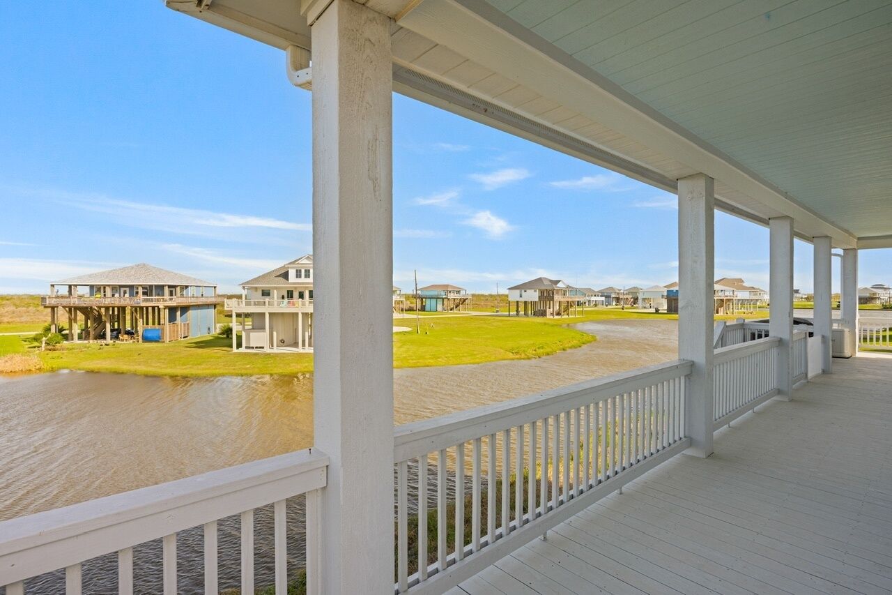 A covered waterfront area with a wooden deck and white railings, overlooking a body of water and several houses.