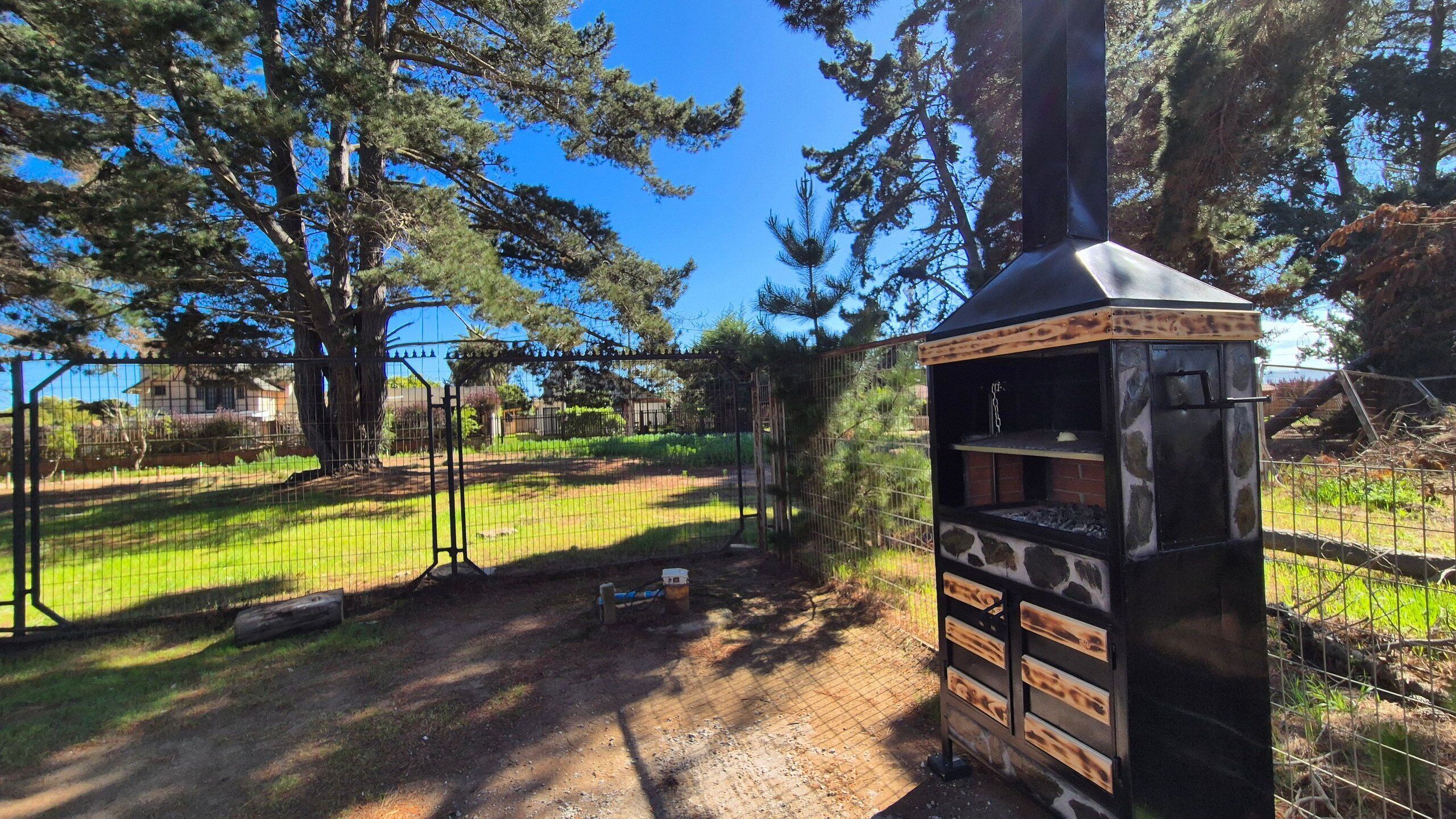 Een rustieke buitenbarbecue met een stenen gevel, omringd door een omheind gebied met bomen en een heldere lucht.