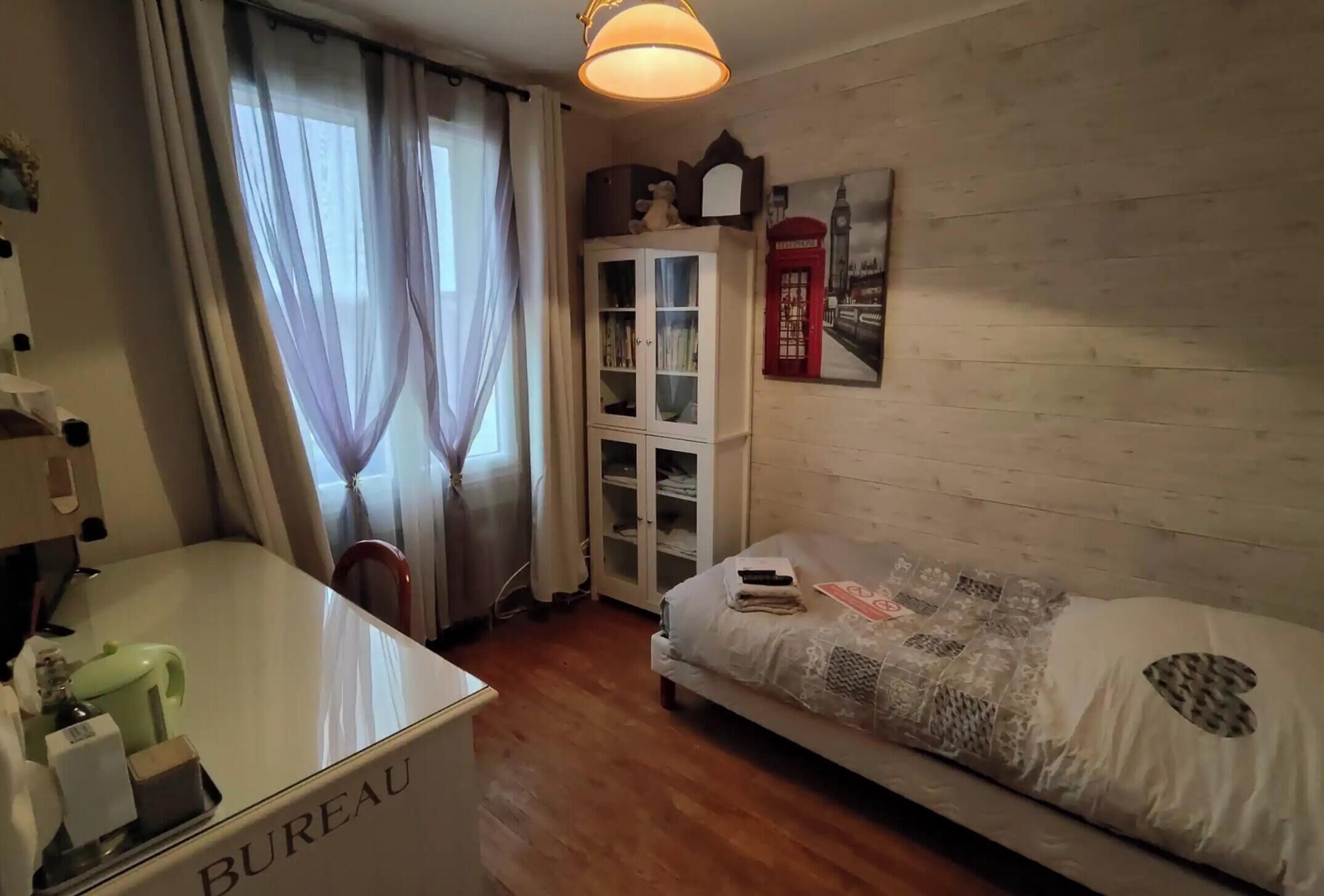 Ein Hotelzimmer mit Schreibtisch, Schrank, Bett, Fenster mit Vorhängen und einem Wandbild.