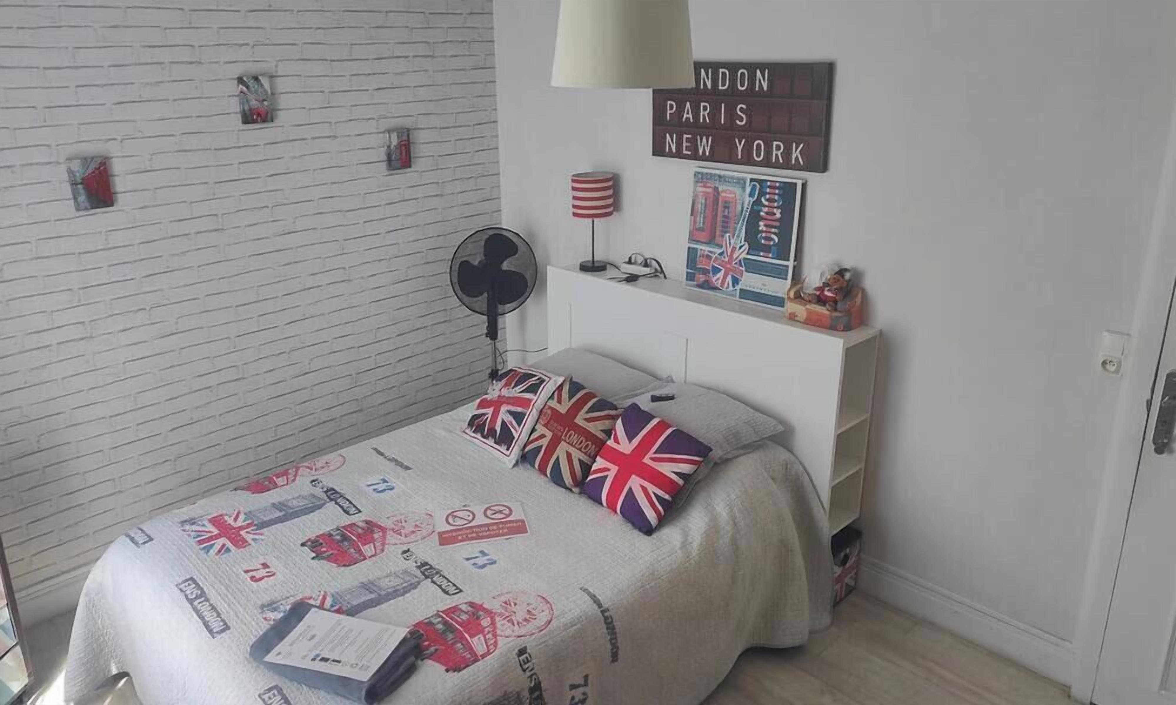 A bedroom with a bed, a white brick wall, a fan, a lamp, and a sign with city names.