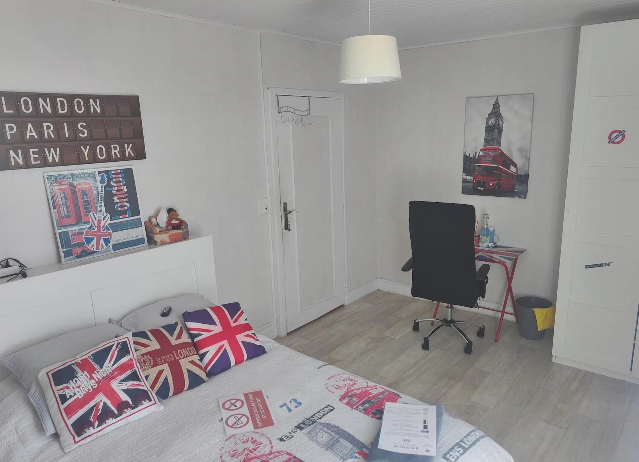 Een slaapkamer met een bed, een bureau, een stoel, een lamp, een deur en een muur met een afbeelding van de London Eye.